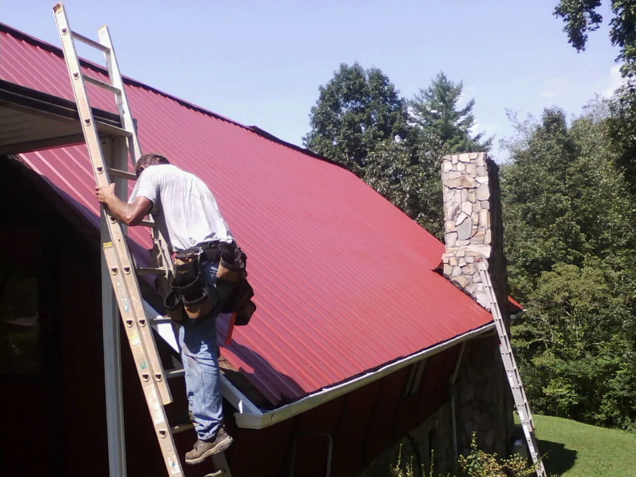 Roofer on a ladder beside a red metal roof and stone chimney — Soffit & Fascia Repair by Roofing Brothers in Polkton